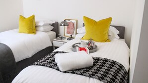 A hotel room with two beds, each with white bedding and yellow pillows. There is a nightstand with a lamp, a framed picture, and a tray with a cup and a thermos.