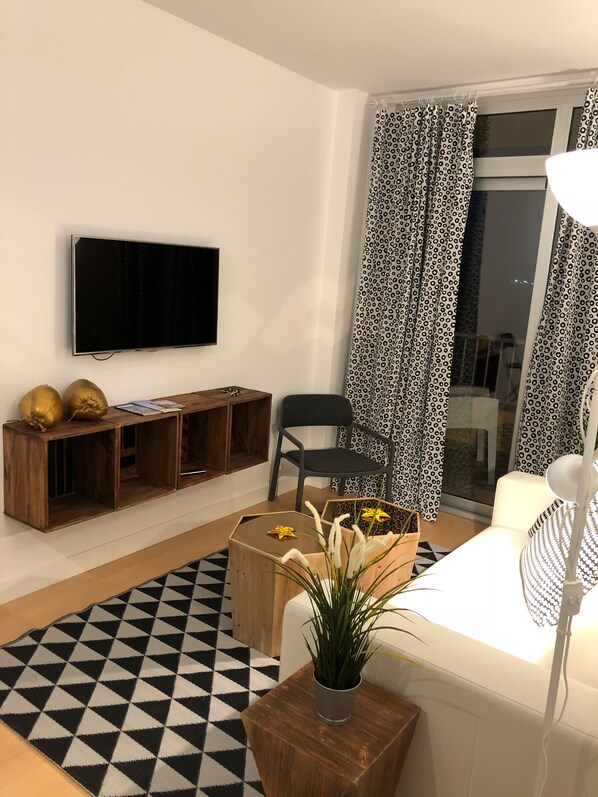 Una sala de estar moderna con televisor de pantalla plana, estanterías de madera, alfombra con diseño blanco y negro, sofá blanco y una planta en maceta.