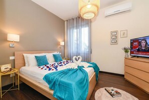 A hotel room with a bed, a television, a small table with a tray of chocolates, and a decorative heart-shaped towel on the bed.