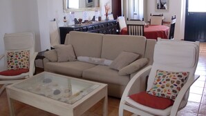 Una sala de estar con un sofá color beige, dos sillones blancos, una mesa de centro de vidrio y un comedor al fondo.