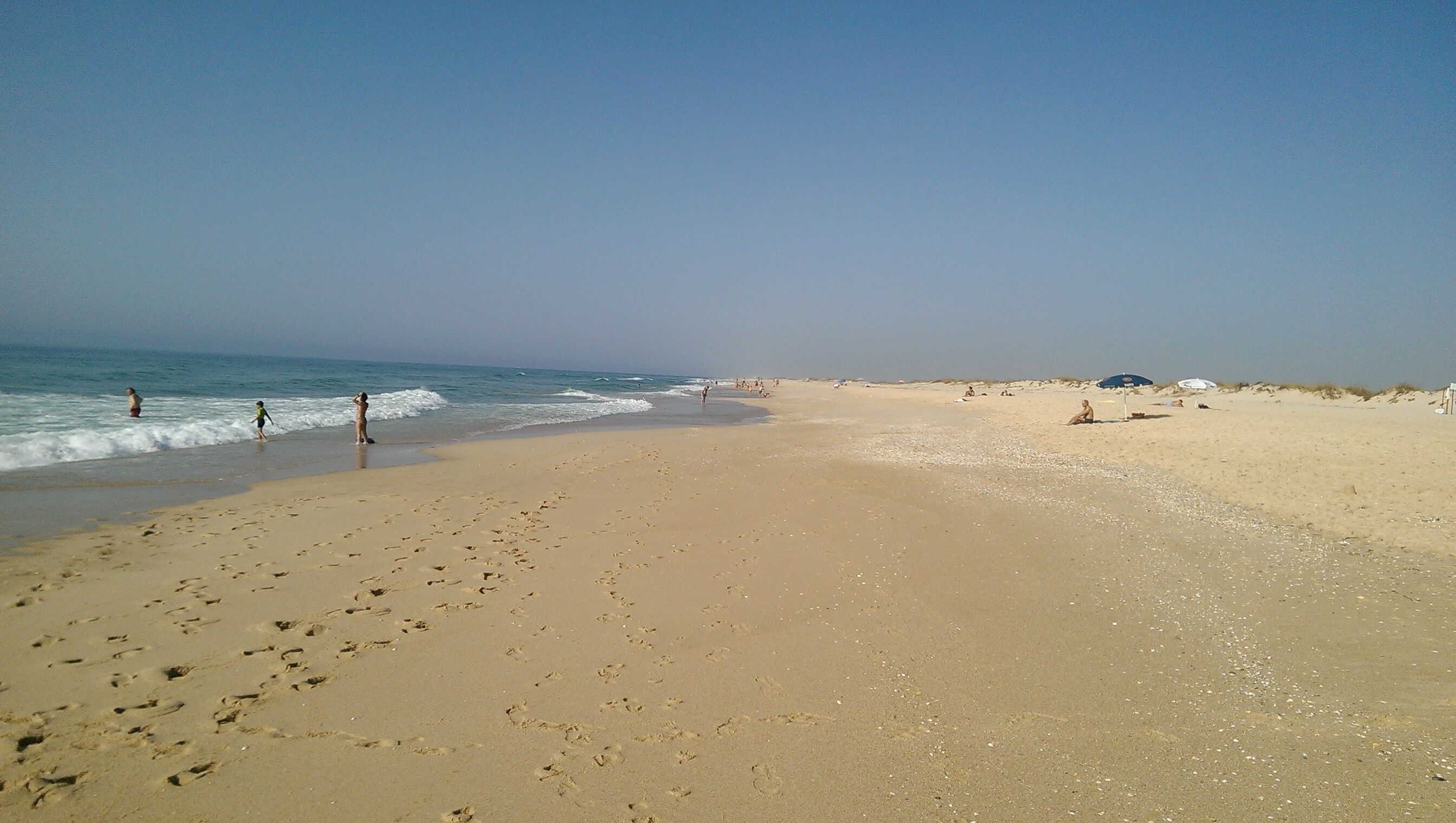 Ein weiter Sandstrand mit Fußspuren, die zum Wasser führen, einige Personen in der Ferne und ein klarer Himmel.