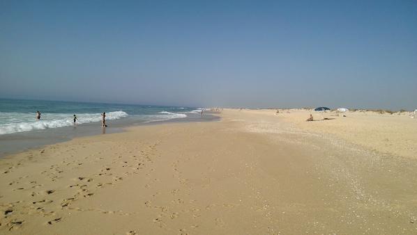 A wide sandy beach with footprints leading to the water, a few people in the distance, and a clear sky. - Casa Magaída, AC, with terrace, near the Ria (Luz (Luz de Tavira))