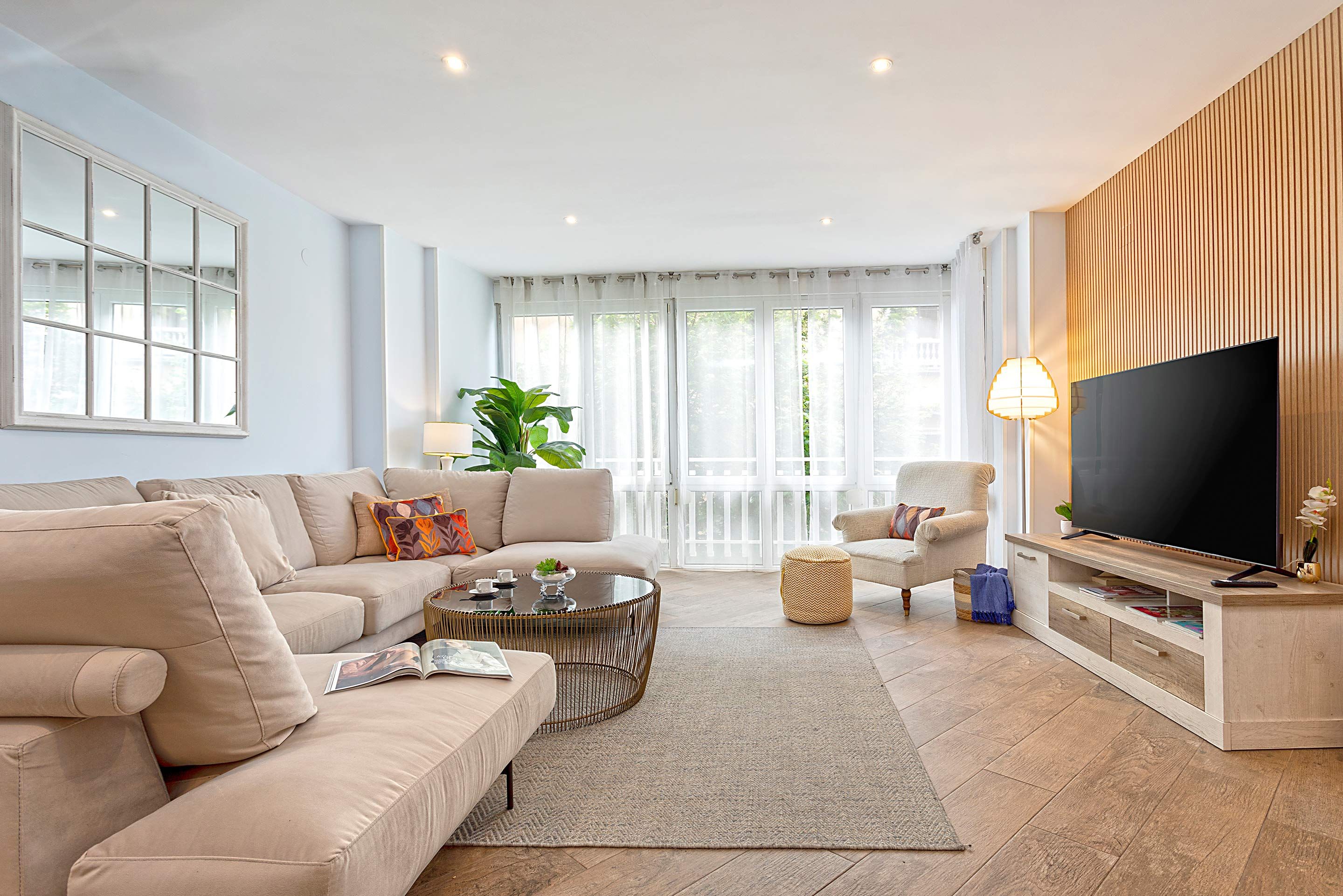 Una sala de estar moderna con un sofá color beige, una mesa redonda, un mueble para televisión y un ventanal con cortinas.