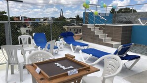A poolside area with lounge chairs, a table with dominoes, and a view of buildings and a church in the background. - Higuey Center City, George City (Higuey)