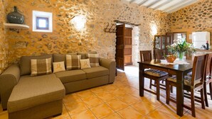 A cozy living room with a stone wall, a sofa, a dining table with chairs, and a kitchen in the background.