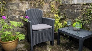 Una silla de mimbre con un cojín, una planta en maceta y una mesita con dos lámparas en un jardín.