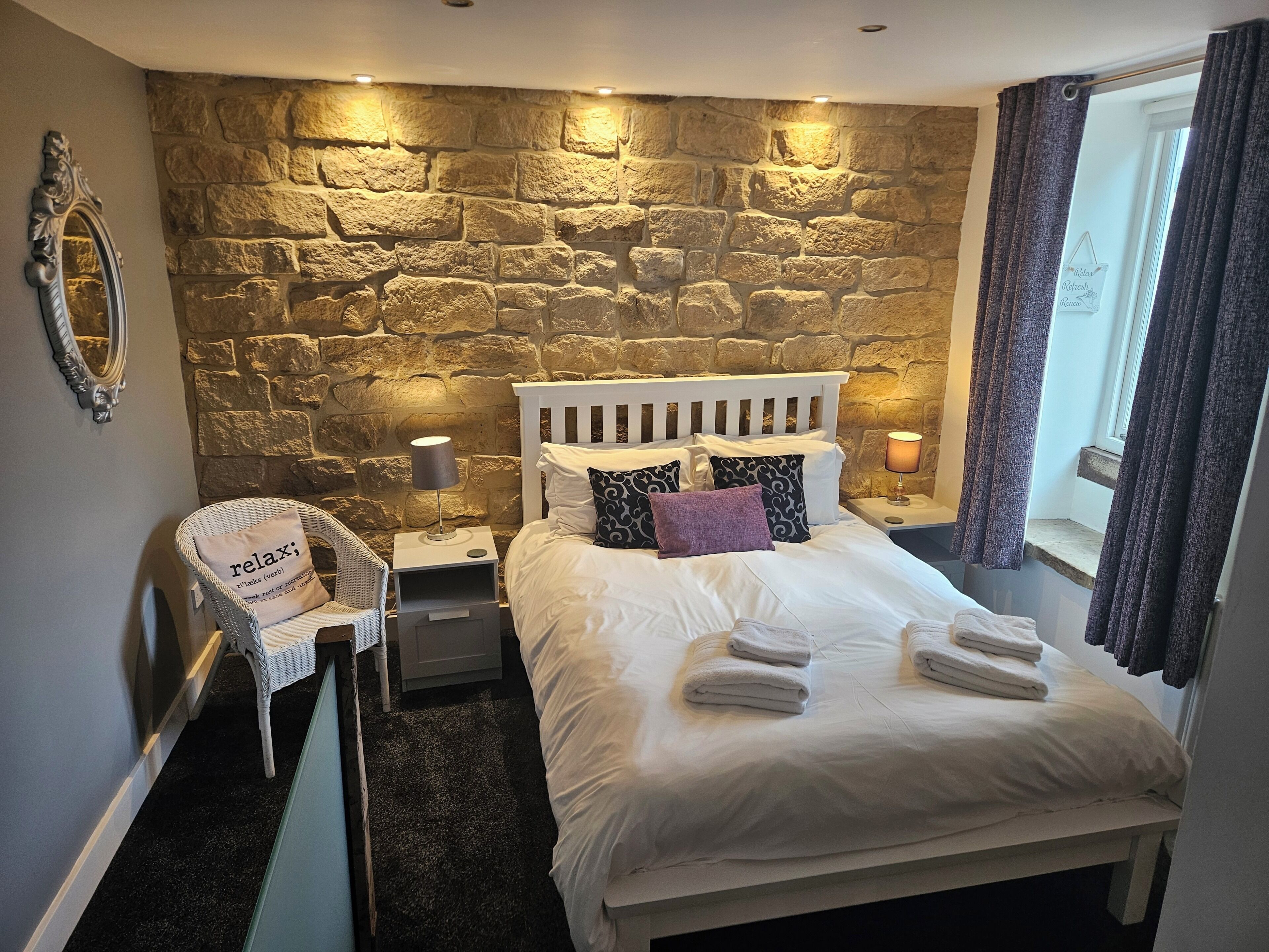 A bedroom with a stone wall, a white bed, a chair, and a window with curtains.