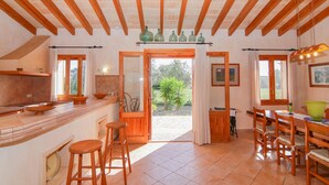 A spacious kitchen with wooden beams, a dining area with wooden chairs, and a view through the open door to the outside.