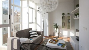 A modern living room with a grey sofa, a black chair, a coffee table with a tray of fruits and a book, a white dandelion sculpture, and a view of the city through large windows. - Singular Stays Botanico 201 (València)