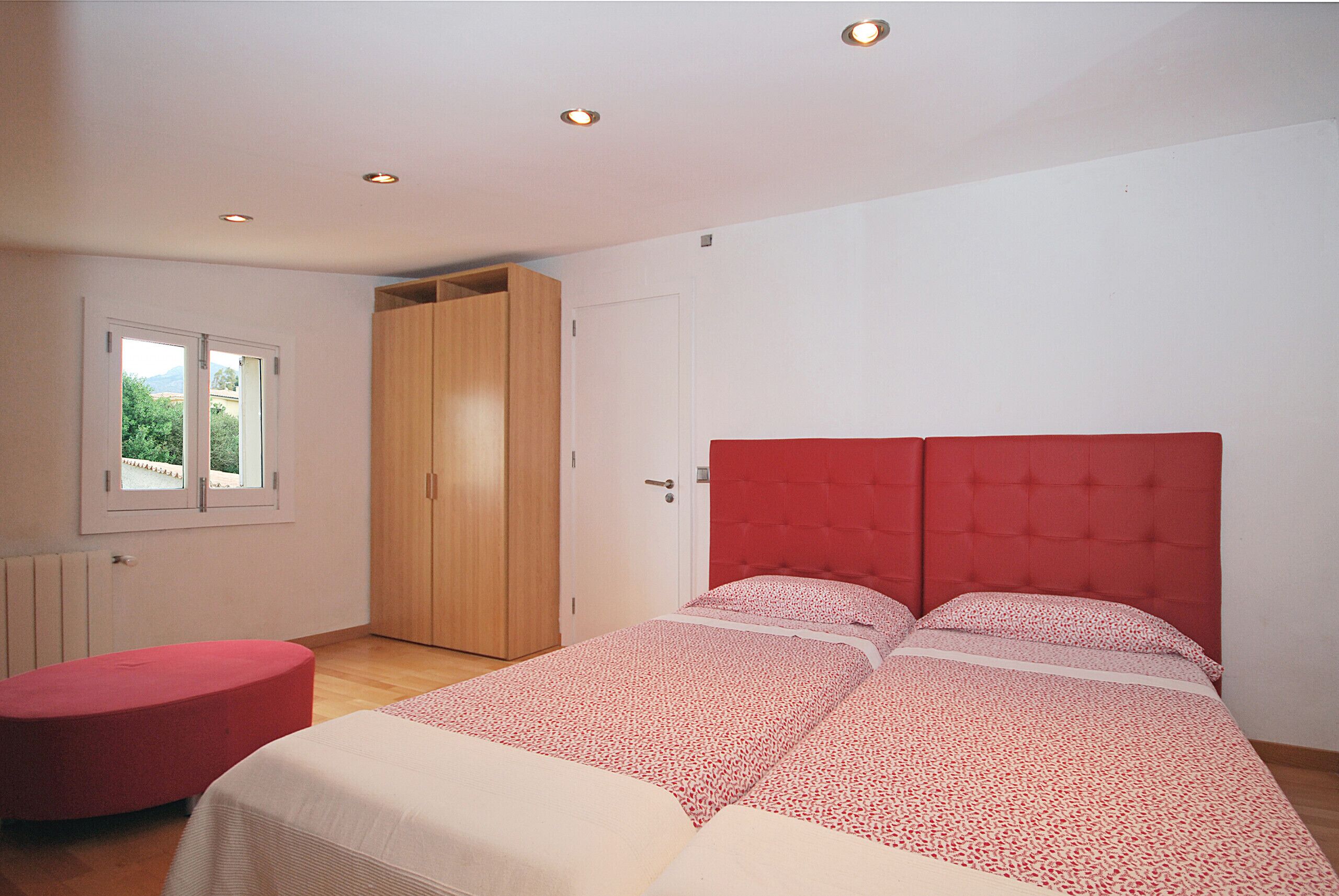 Ein Schlafzimmer mit einem Bett, einem roten Pouf, einem Kleiderschrank und einem Fenster.