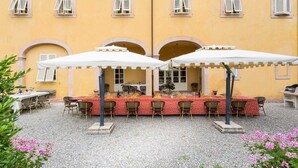 An outdoor dining area with tables and chairs, covered by white umbrellas, set against a yellow building with arched doorways. - Villa Controni     . (Capannori)