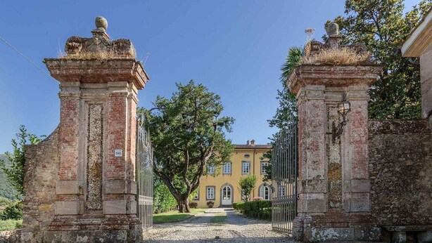 An entrance with two stone pillars, a cobblestone path, and a building in the background. - Villa Controni     . (Capannori)