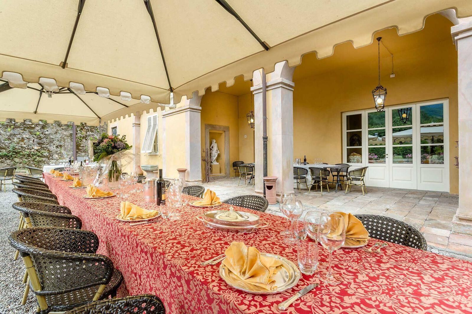 A long dining table set for a meal with wicker chairs, a red tablecloth, and a garden view.
