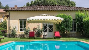 A swimming pool with two red chairs and a white umbrella in front of a building with a tiled roof. - Villa Controni     . (Capannori)
