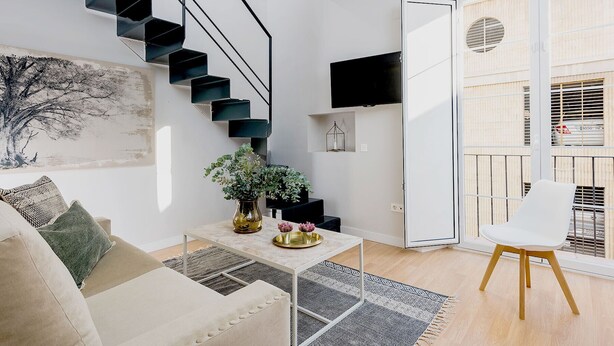 A modern living room with a staircase, a flat-screen TV, a white chair, and a sofa.