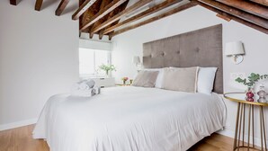 A bedroom with a large bed, wooden beams, a bedside table with a lamp, and a small round table with decorative items.