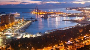 A coastal cityscape at dusk with illuminated buildings, a port with cranes, and a bridge.