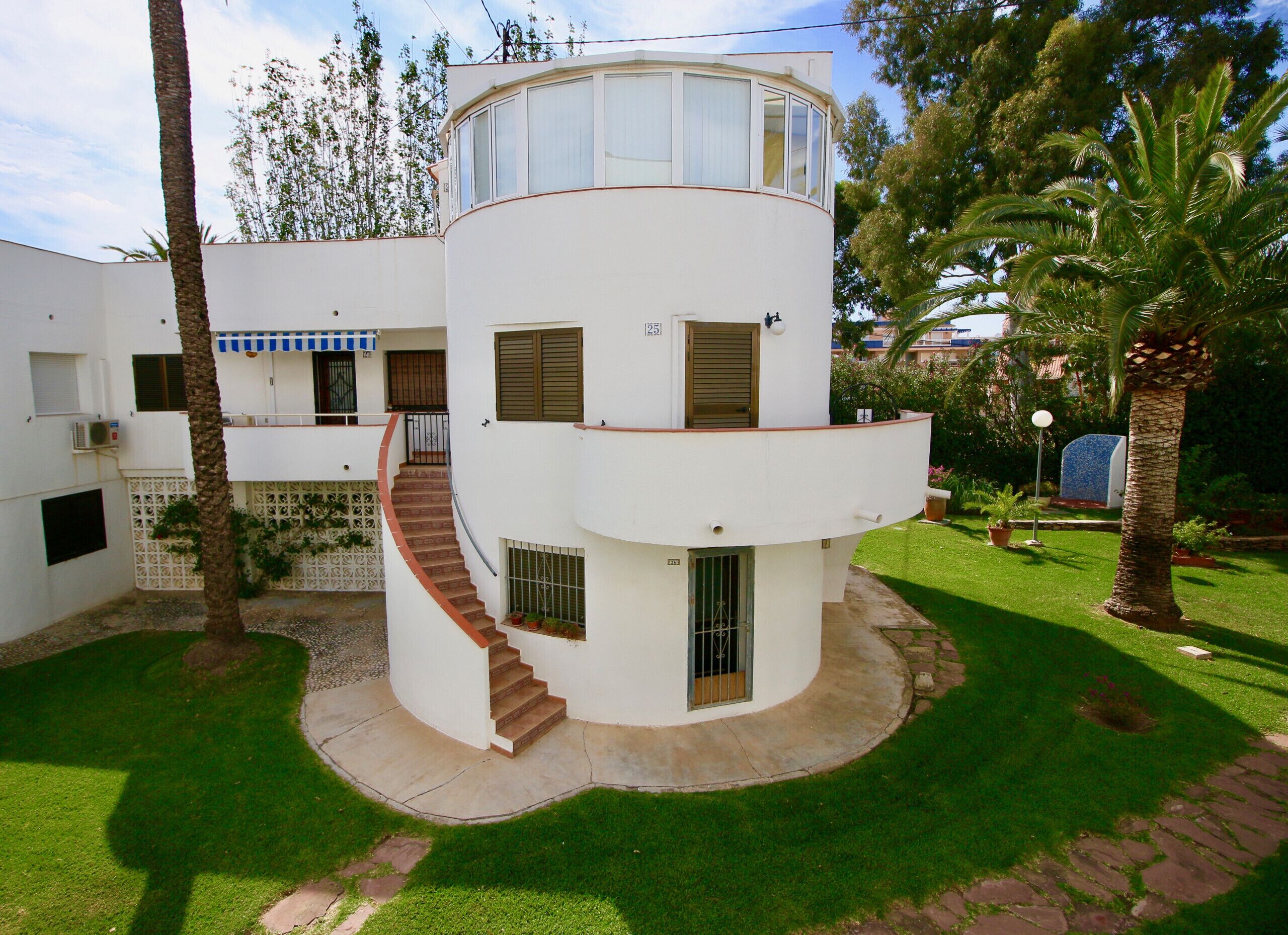 A two-story building with a curved staircase, a balcony, and a small garden area with palm trees.