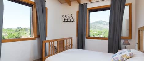 A bedroom with a bed, two windows with curtains, a wooden ceiling, a wall-mounted light, and a view of greenery outside.