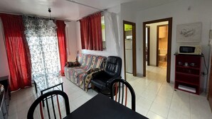A living room with a sofa, a dining table with chairs, a TV, and a kitchen area visible through an open doorway. - Siroco Holidays 5-B LEK (Peñiscola)
