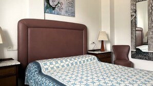 A hotel room with a brown headboard, a patterned bedspread, a nightstand with a lamp, a chair, a mirror, and a painting of roses on the wall. - Siroco Holidays 5-B LEK (Peñiscola)