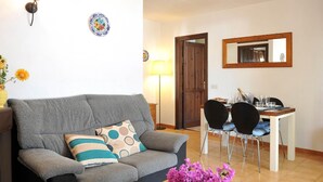 Una sala de estar con un sofá gris, una mesa de centro con un florero, una zona de comedor con una mesa y sillas, y una puerta de madera.