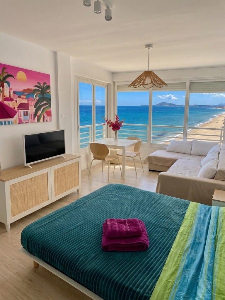 Une chambre à coucher comprenant un lit, une télévision, une table à manger et un canapé. Une grande fenêtre offre une vue sur la plage.