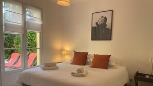 En snyggt bäddad säng med vita lakan och orange kuddar, ett sängbord med en lampa och en inramad tavla på väggen.