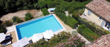 A rectangular swimming pool surrounded by lush greenery and a white building with a tiled roof.