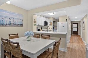 A modern kitchen with white cabinets, a central island, and a dining area with wicker chairs and a vase of flowers. - CRC 1303 - Silver Oceanfront Extended Balcony (St Augustine)