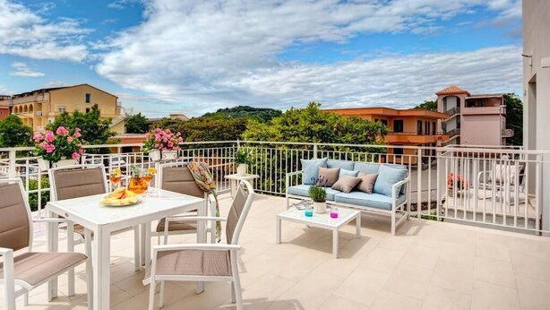 Ein Balkon mit weißem Tisch, Stühlen und einer Couch. Übertöpfe mit Pflanzen und ein Geländer sind ebenfalls vorhanden.