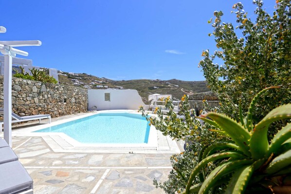 Zona de piscina con suelo de piedra, sillas de descanso blancas y vistas a una colina con edificios.