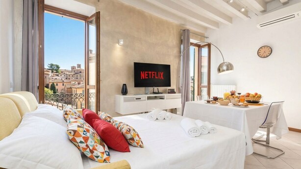 A hotel room with a bed, a TV displaying 'NETFLIX,' a dining table with breakfast items, and a view of buildings outside. - DesenzanoLoft Le Petit Bijou - Restyling 2025 - Downtown (Desenzano del Garda)