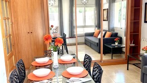 Un área de comedor moderna con una mesa de vidrio, sillas negras y manteles individuales anaranjados. Se ve una puerta de madera y un sofá al fondo.