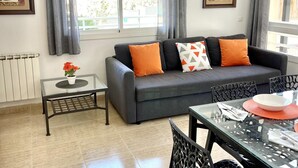 Una sala de estar moderna con un sofá gris, cojines naranjas, una mesa de centro de vidrio y un comedor con mesa y sillas de vidrio.