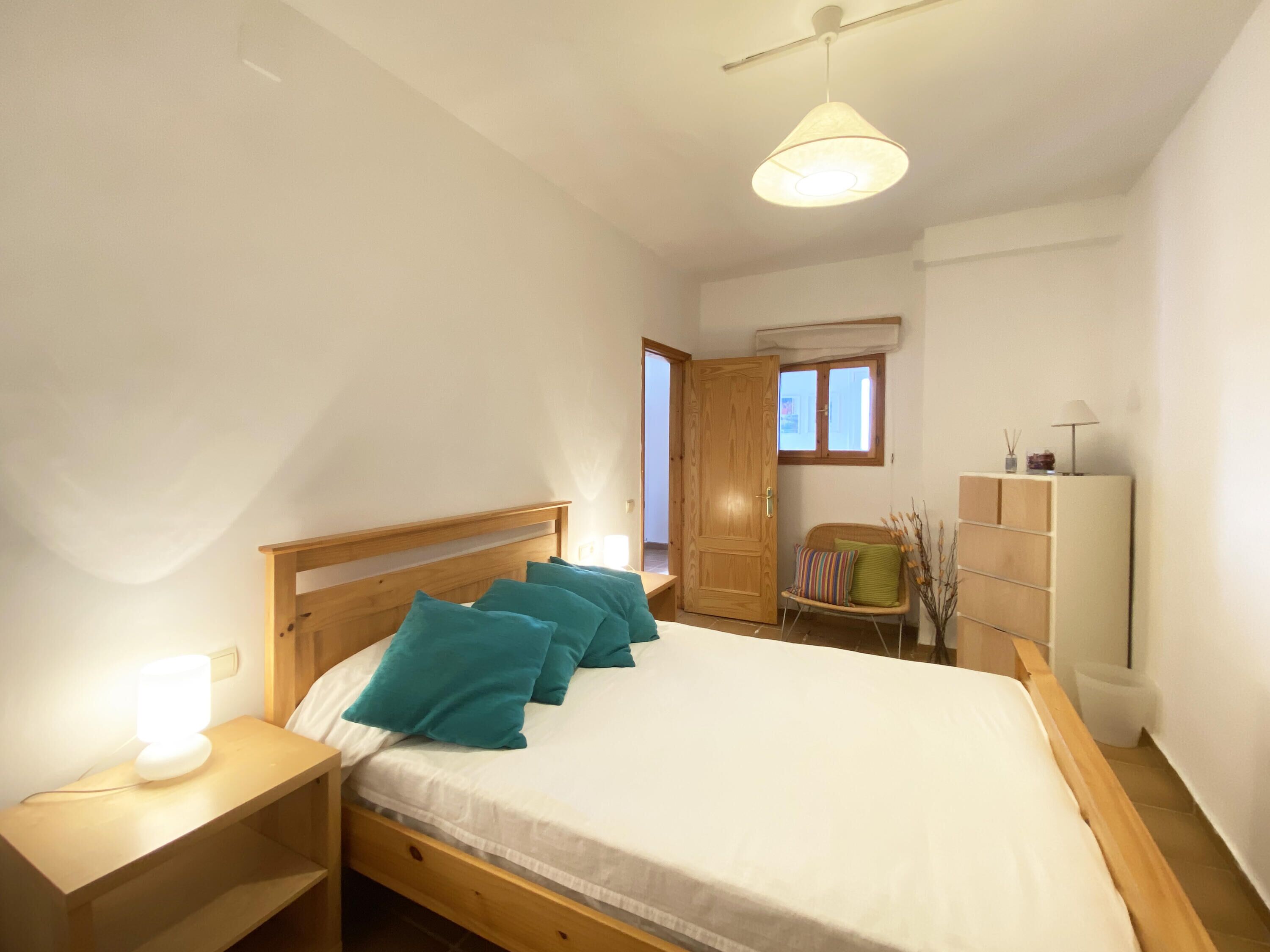 Quarto com cama de madeira, almofadas verde-água, mesa de cabeceira com abajur, guarda-roupa e janela com vista.