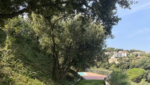 A pathway leading to a pool surrounded by lush greenery and trees, with a view of houses in the background. - Holiday rental semidetached with swimming pool in Begur, Sa Riera (Begur)