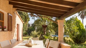 A covered patio with a table and chairs, surrounded by lush greenery and a view of the garden. - Casa Tonina - Beautiful villa with pool and garden in Pollença (Pollença)