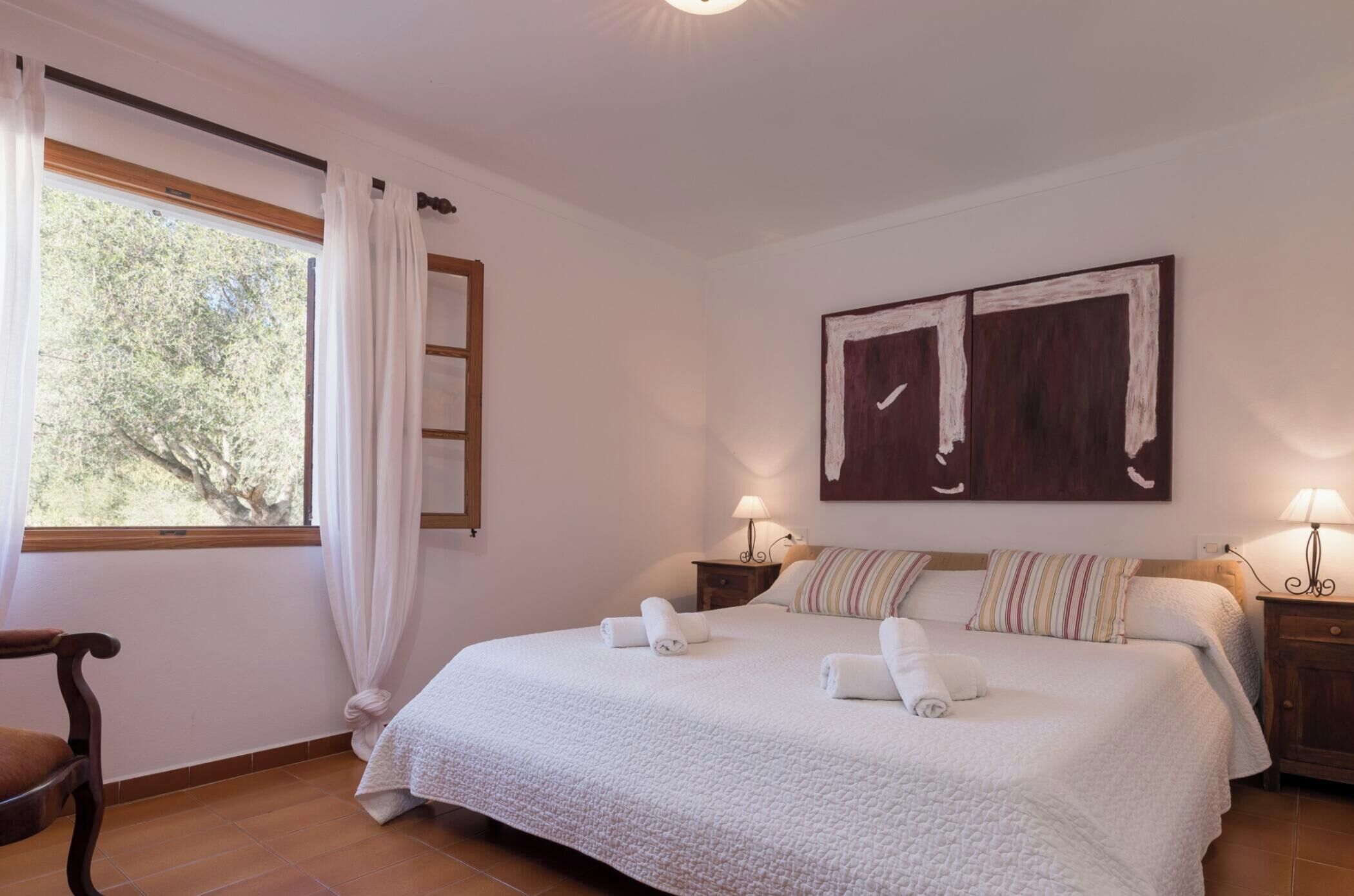 A bedroom with a large bed, two bedside tables with lamps, a wooden chair, a window with curtains, and a painting on the wall.