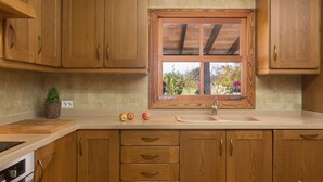 A kitchen with wooden cabinets, a window with a view of greenery, a sink, and a countertop with apples. - Casa Tonina - Beautiful villa with pool and garden in Pollença (Pollença)