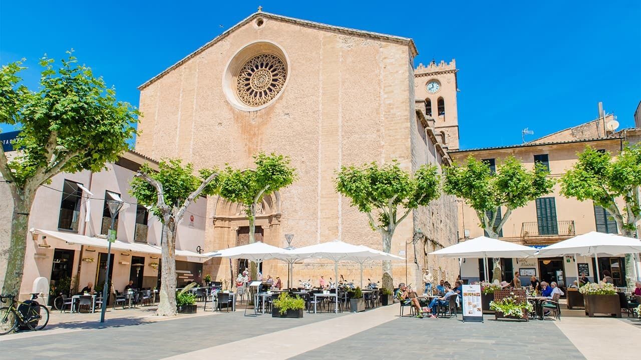 An outdoor café area with tables and umbrellas in front of a historic building with a large circular window and a clock tower.