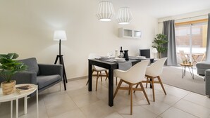 Un salon moderne avec un canapé, une table à manger et des chaises. On y trouve une lampe, une plante en pot et une petite table sur laquelle est posé un livre. La pièce est dotée d’une grande fenêtre avec des rideaux.