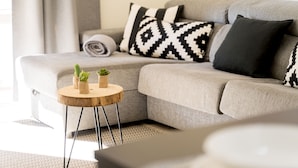 Un salon moderne avec un canapé d’angle gris, deux coussins aux motifs noir et blanc et une petite table d’appoint en bois sur laquelle sont posées deux plantes en pot.