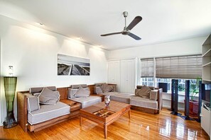 A modern living room with a wooden floor, a ceiling fan, a large window with blinds, and a painting on the wall.
