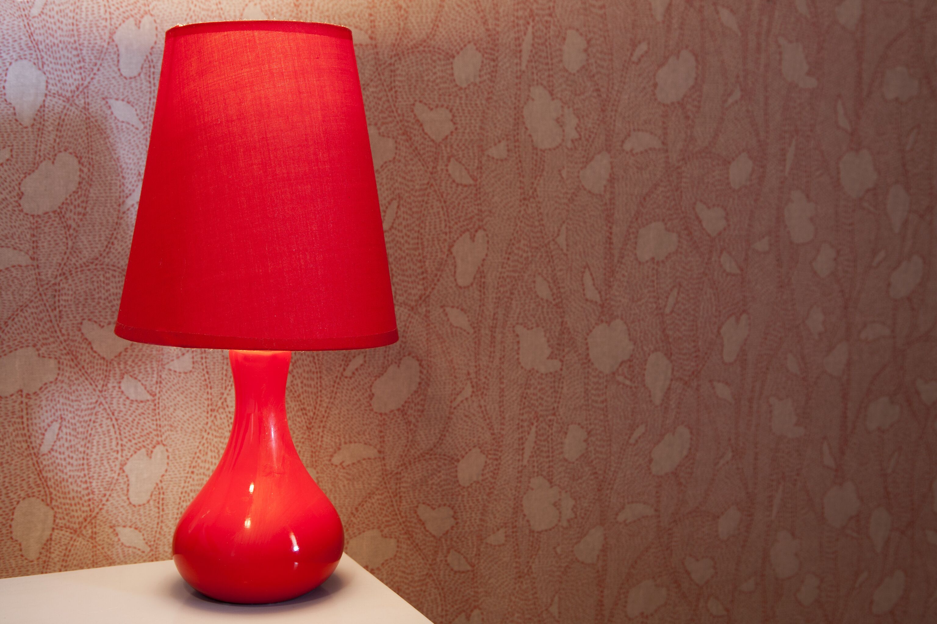 A red table lamp on a white surface against a patterned wall.