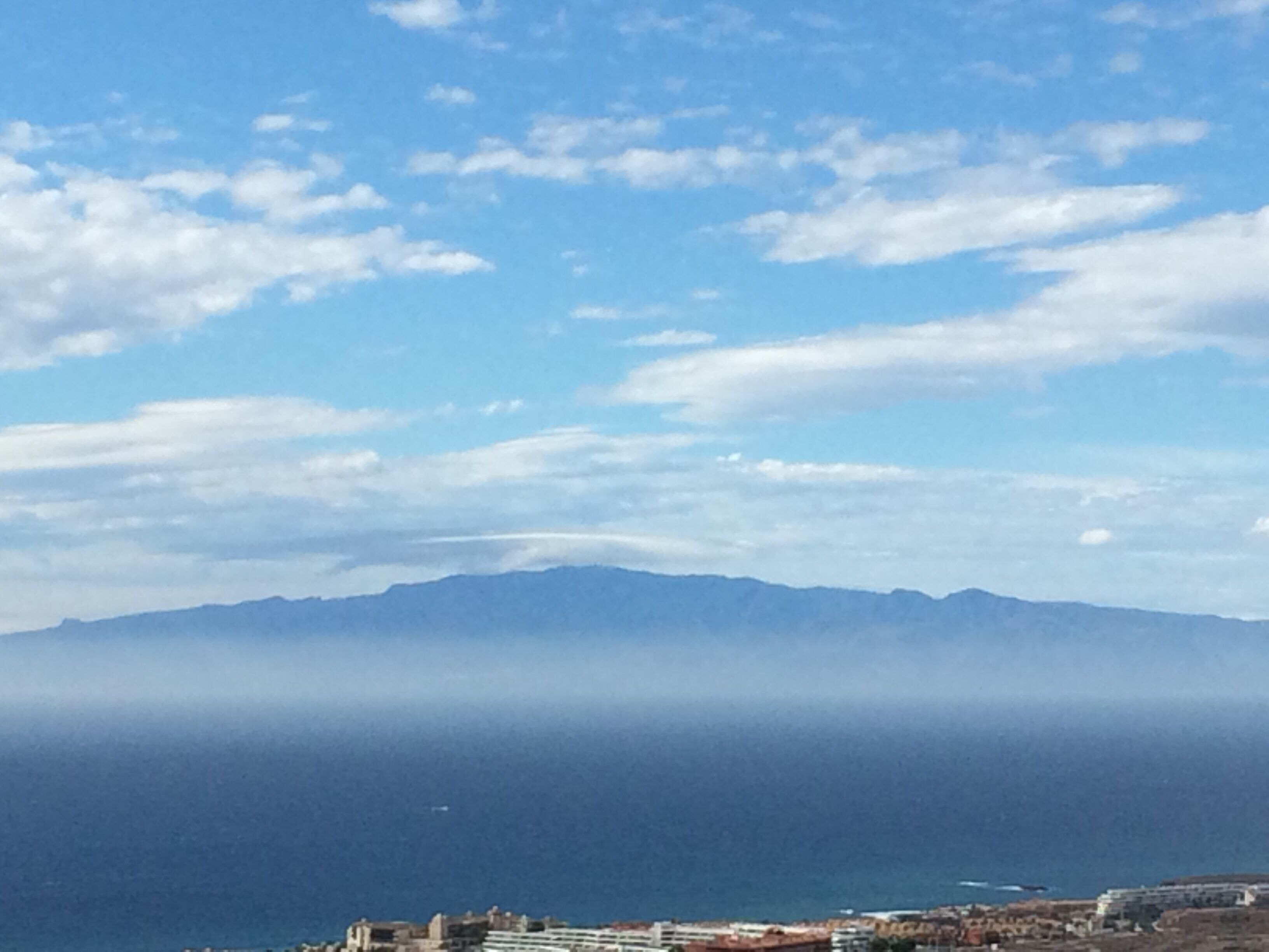 Un pueblo costero con edificios cerca del mar, una montaña al fondo y un cielo despejado con nubes.