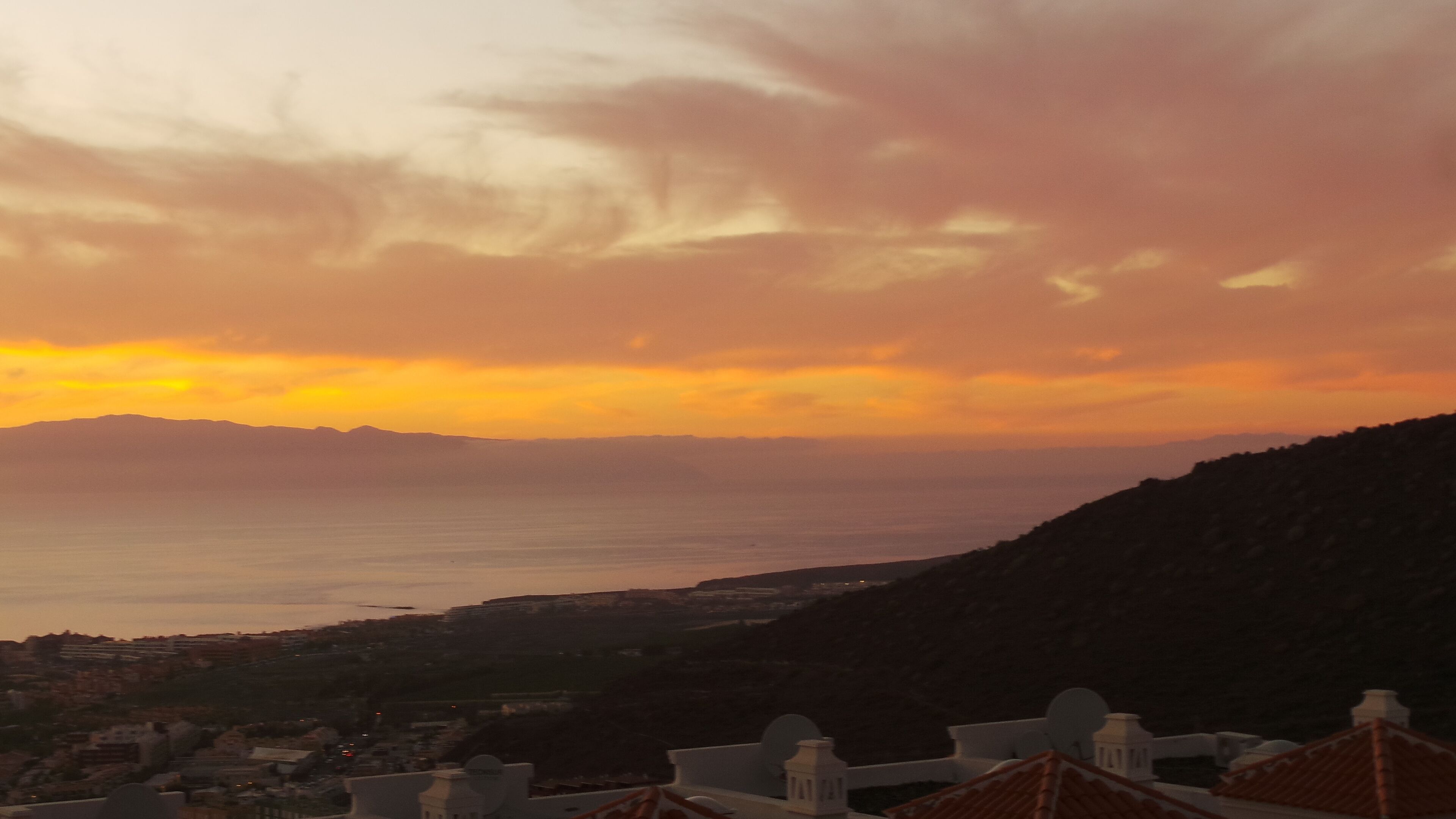 Paisagem costeira com pôr do sol, montanhas e uma cidade abaixo.