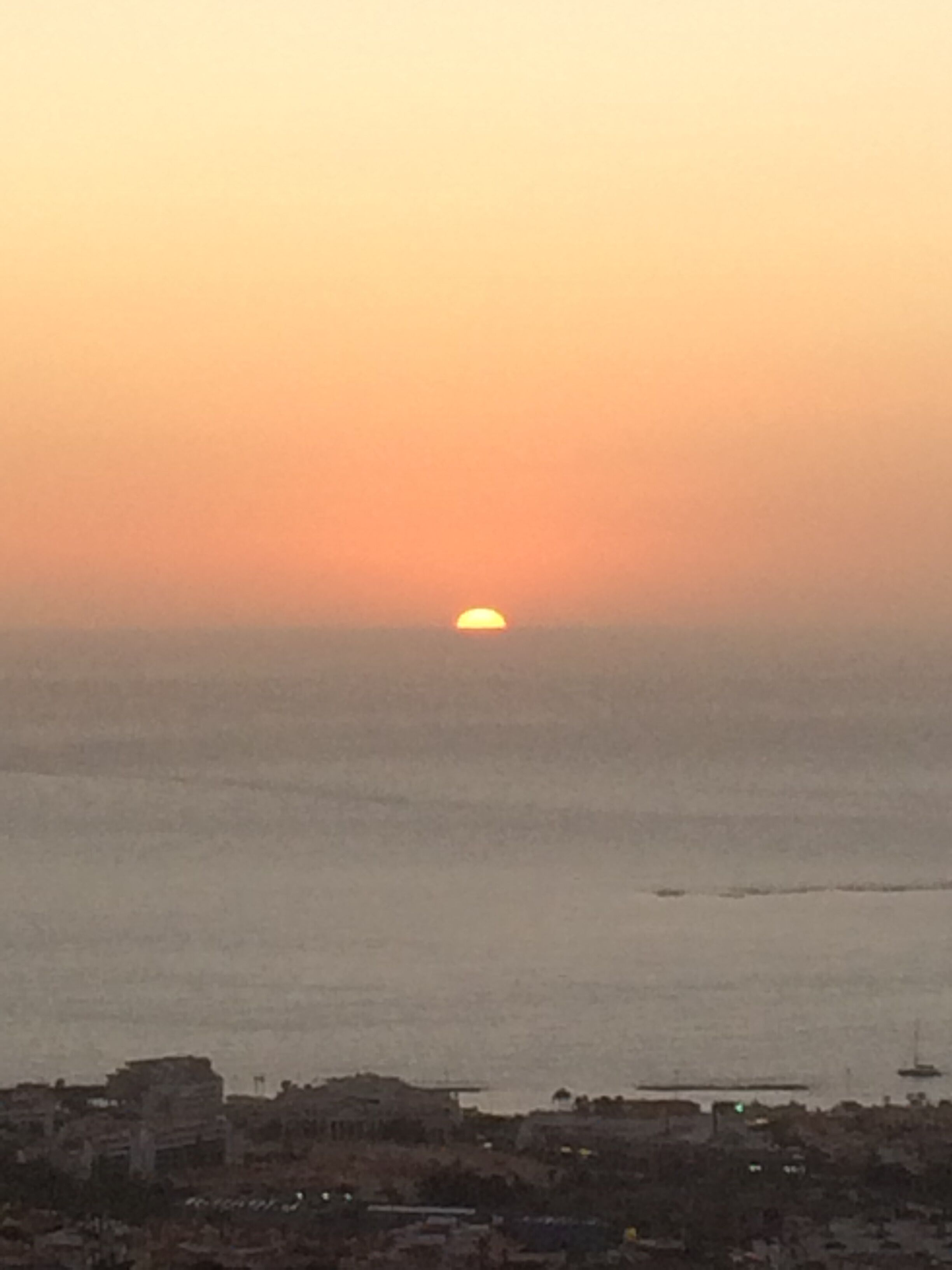 A coastal cityscape at sunset with a visible sun near the horizon.