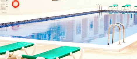 A swimming pool with two green lounge chairs, a lifebuoy, and a tiled border.