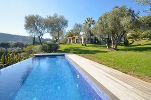 Une piscine dotée d’une terrasse en bois, entourée d’une végétation luxuriante, avec une maison en arrière-plan.